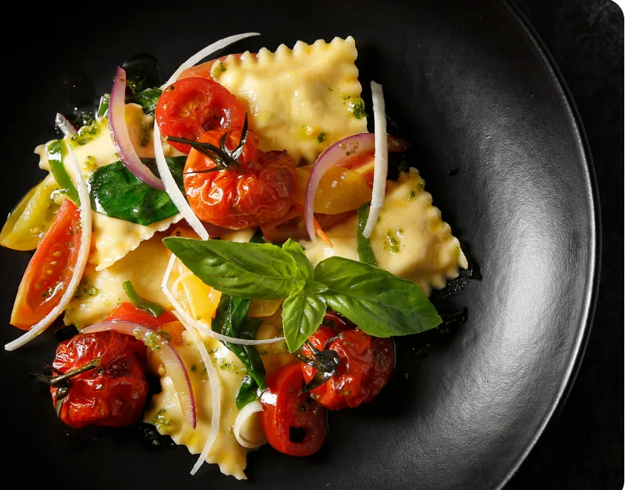 A close-up overhead shot of a plated pasta dish in a black bowl. The dish appears to be ravioli with roasted red tomatoes, sliced red onion, green herbs, and a basil leaf on a dark background.